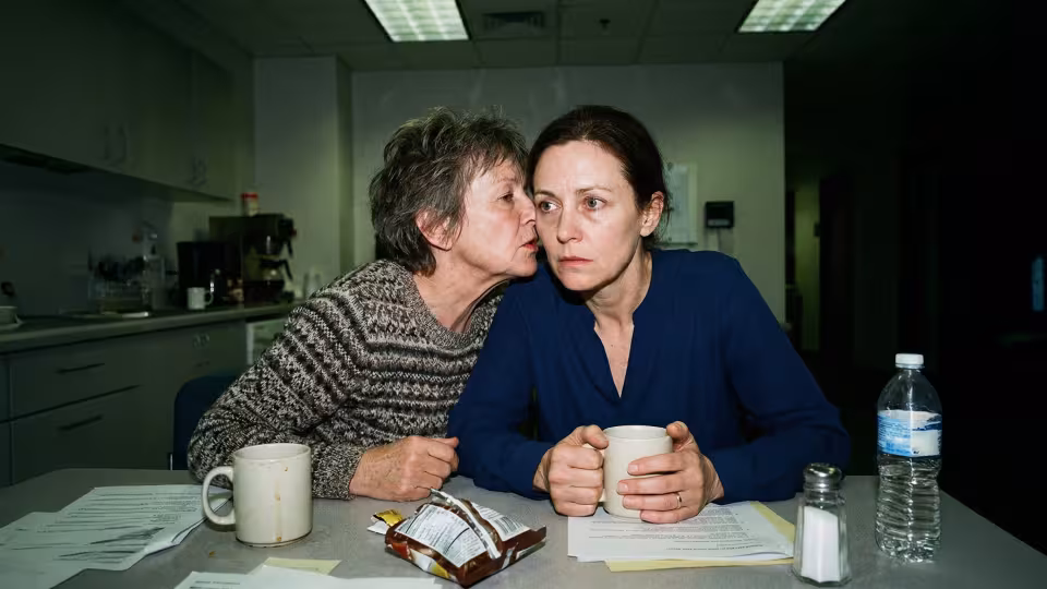 Dvě ženy se spiklenecky naklánějí nad levný laminátový stůl v ostré kancelářské kuchyni osvětlené zářivkami.