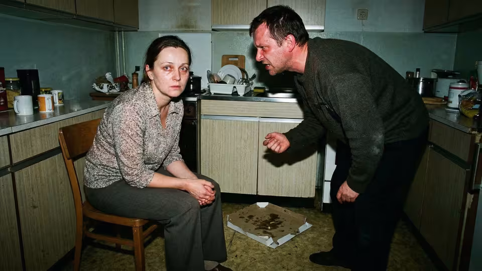 Žena sedí na obyčejné kuchyňské židli, její tělo je mírně shrbené, zatímco muž stojí nad ní, s tváří zkroucenou směsicí vzteku a zoufalství.