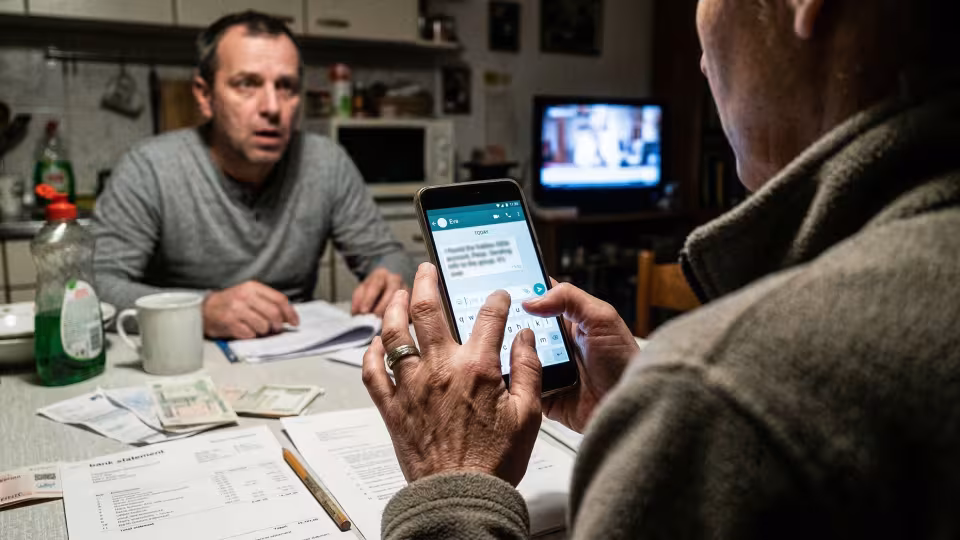 Nízkoúhlý, trochu mimo mísu záběr zpoza ramena hlavní hrdinky, při pohledu na obrazovku svého smartphonu, jasně osvětlenou proti zšeřelé, přeplněné kuchyni.