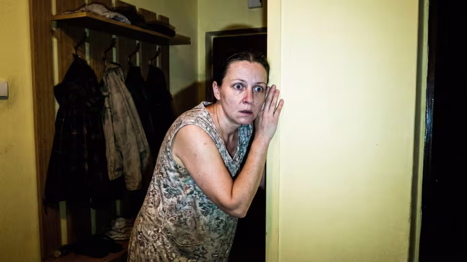 In a dimly lit hallway of a contemporary Prague panelák apartment, a woman's face is caught in a harsh, direct camera flash, revealing wide eyes filled with shock and dawning horror. Her body is frozen in a posture of intense, desperate listening, ear pressed tightly to a thin, pale yellow wall.