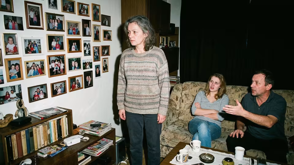 Uprostřed stojí žena s pohledem upřeným na hustě zaplněnou stěnu zarámovaných rodinných fotografií