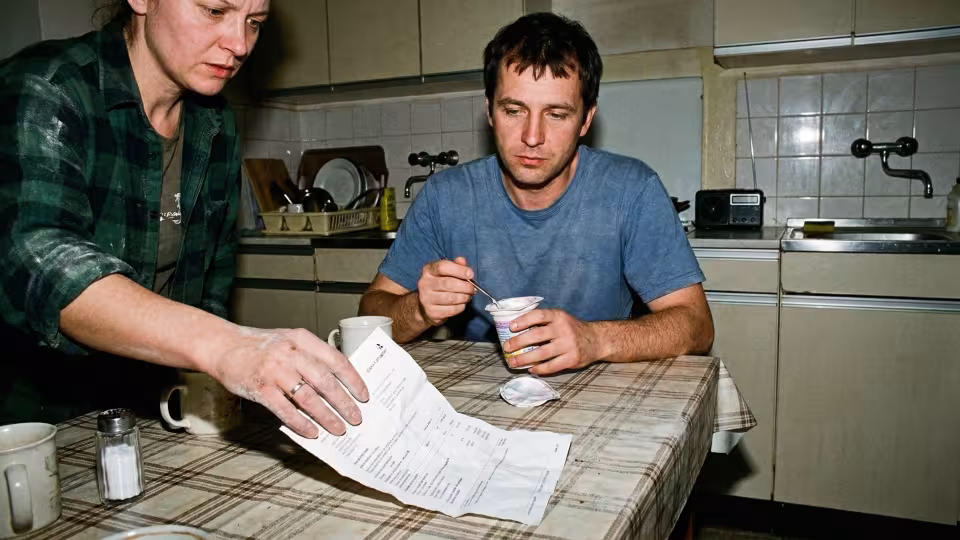 A woman's left hand, visibly smudged with fine construction dust, trembles as it releases a crumpled bank statement onto the surface.