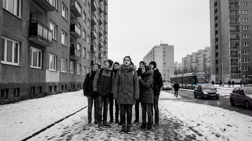Širokoúhlý záběr z nízkého úhlu zachycuje skupinku teenagerů na zasněžené betonové cestě mezi dvěma holskými panelákovými bloky.