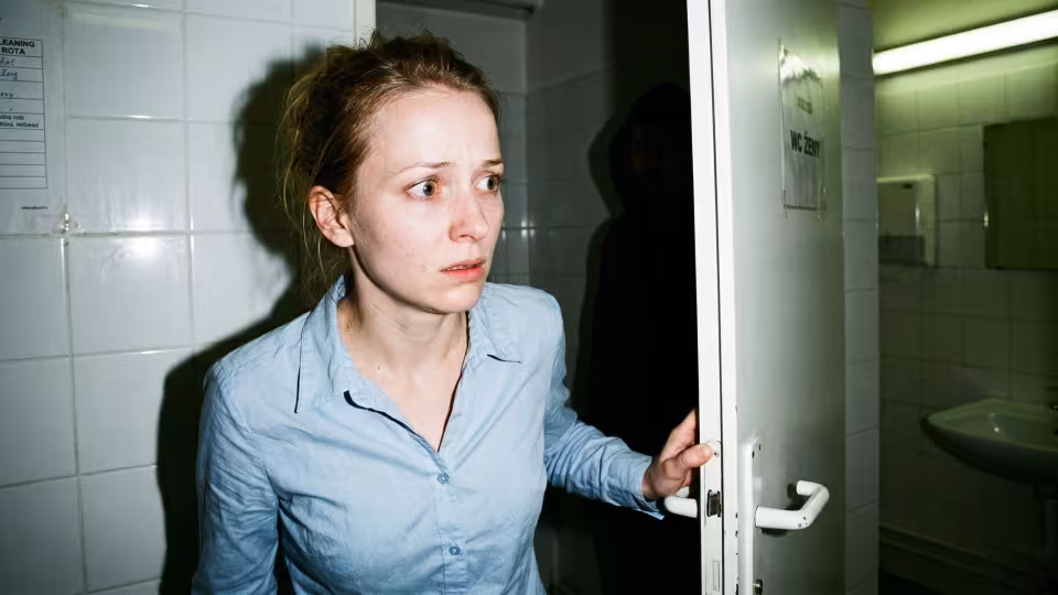Úzký, trochu nekonvenční záběr šestadvacetileté ženy s tváří bledou s nastupující hrůzou, jednou rukou stále svírající bílou, úřední kliku toaletní kabinky.