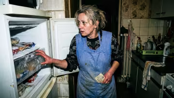 Nízkoúhlý, těsný záběr ženy ve stísněné, datované české panelákové kuchyni. Její záda jsou strnule přitisknutá k chladnému, bílému povrchu starého mrazáku, tělo zmrzlé.