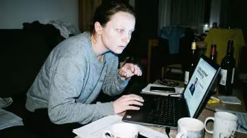 A woman is hunched over a laptop in a dimly lit, cluttered living room of a contemporary Czech panelák apartment. Her eyes are wide with shock, reflecting the harsh glow of the laptop screen.
