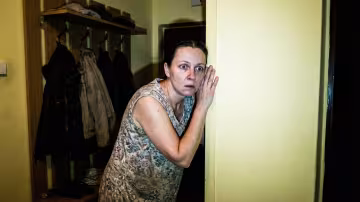 In a dimly lit hallway of a contemporary Prague panelák apartment, a woman's face is caught in a harsh, direct camera flash, revealing wide eyes filled with shock and dawning horror. Her body is frozen in a posture of intense, desperate listening, ear pressed tightly to a thin, pale yellow wall.