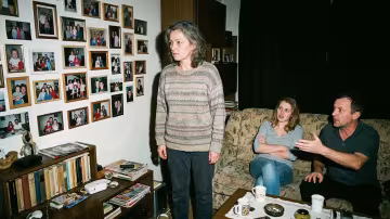 Uprostřed stojí žena s pohledem upřeným na hustě zaplněnou stěnu zarámovaných rodinných fotografií